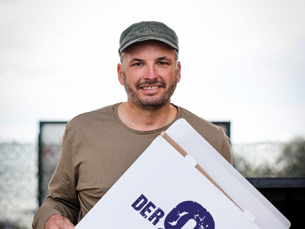 Zu sehen ist: der Kabarettist HP Arzberger. Er lächelt in die Kamera, hat eine graue Kappe auf dem Kopf, ein hellbraunes T-Shirt an und hält eine Pizzaschachtel in der Hand.